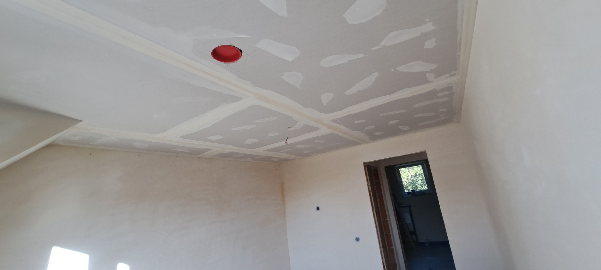 Wnętrze pokoju w trakcie prac wykończeniowych: sufit z płyt kartonowo-gipsowych z widocznymi łączeniami i otworem na oświetlenie w kolorze czerwonym, ściany częściowo pokryte gładzią, widoczny...