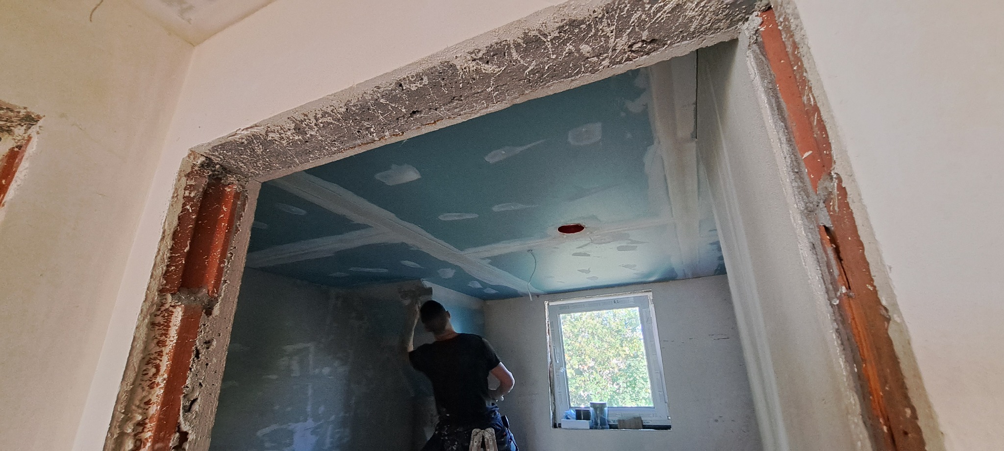 Pracownik szpachluje ścianę w pomieszczeniu z zamontowaną płytą karton-gips na suficie, widoczne surowe wykończenie ościeżnicy drzwiowej.