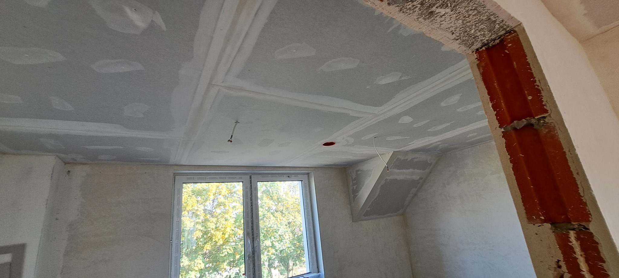 Sufit z płyt kartonowo-gipsowych w trakcie montażu, widoczne łączenia i szpachlowanie, wystające przewody elektryczne, surowy mur przy otworze drzwiowym, okno z widokiem na drzewa.