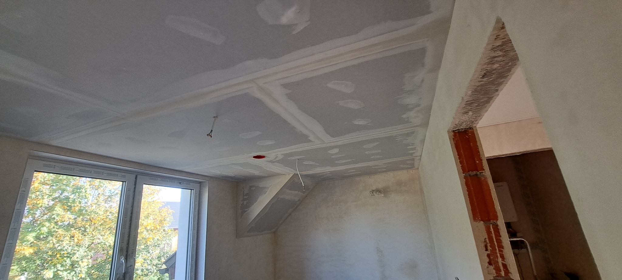 Pokój w trakcie remontu: sufit z płyt kartonowo-gipsowych ze szpachlowanymi łączeniami, widoczne kable elektryczne, okno z widokiem na drzewa, surowa ściana z otwartym przejściem do innego...