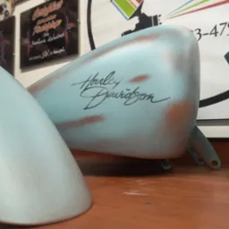 Pomalowane na niebiesko z efektem rdzy elementy motocykla Harley Davidson: błotnik, zbiornik paliwa z napisem i element ramy, leżą na drewnianym blacie.