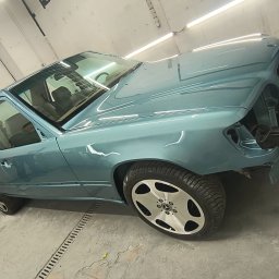 Jarosław Kuśmierek Custom Painting - Turkusowy Mercedes W124 bez przedniego zderzaka w lakierni, po świeżym lakierowaniu. Widoczne odbicia światła na karoserii, podkreślające gładkość powłoki.