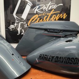 Jarosław Kuśmierek Custom Painting - Szare, polakierowane elementy motocykla Harley-Davidson: błotnik, zbiornik paliwa i kufer, z czarnym napisem i czerwoną linią, na tle plakatu Retro and Custom.