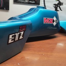 Jarosław Kuśmierek Custom Painting - Turkusowy bak i błotniki motocykla MZ ETZ 250 po renowacji lakierniczej, z naklejkami. Błyszcząca powierzchnia, widoczne odbicia światła. Tło: ściana z grafikami.