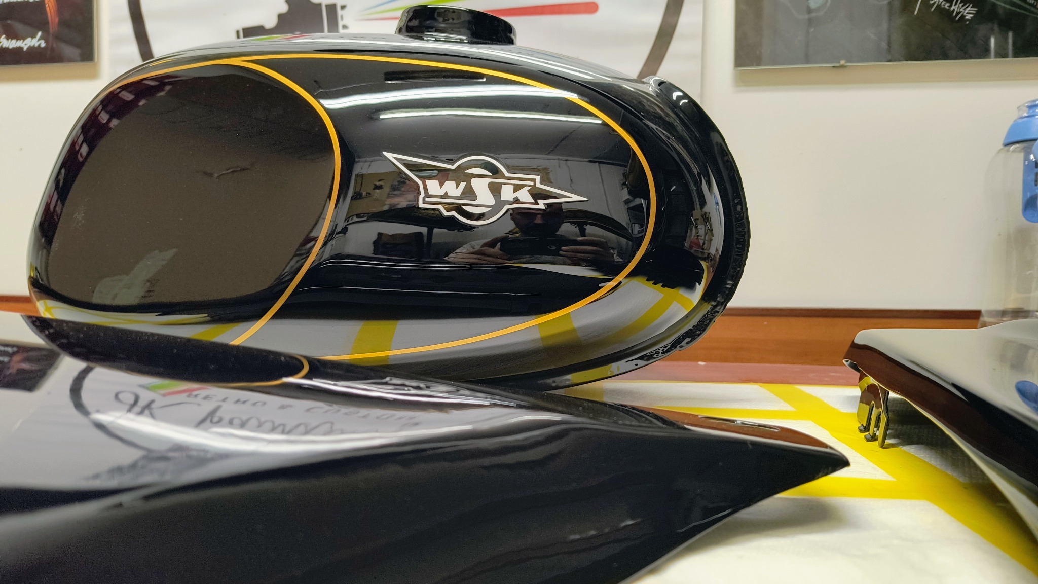 Czarny bak motocyklowy WSK z żółtymi obrysami i srebrnym logo, po renowacji lakierniczej, widoczne odbicie fotografa w lakierze.