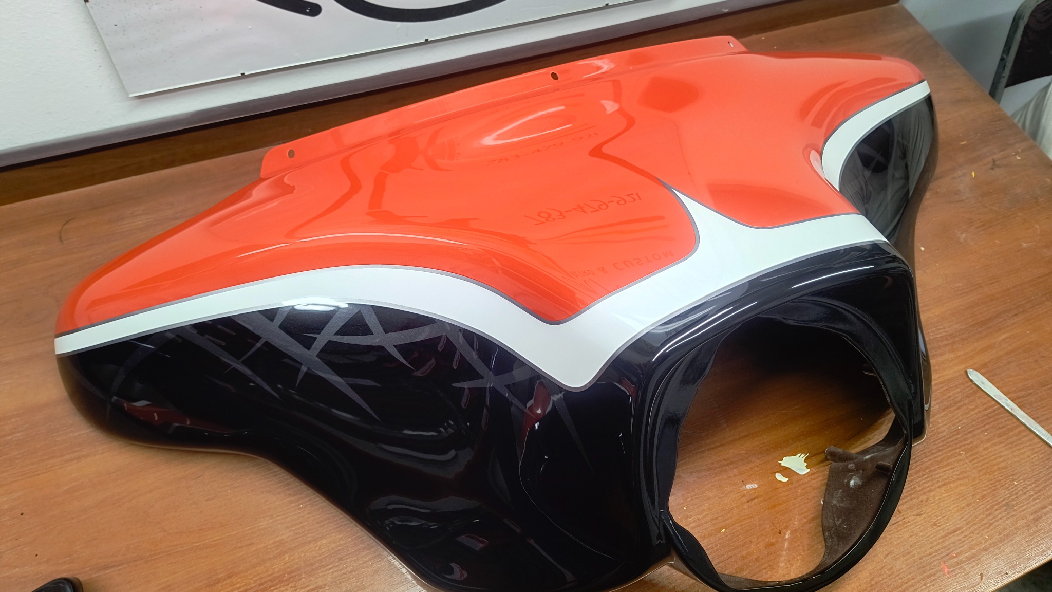 Pomarańczowo-czarna owiewka motocyklowa z białym paskiem i szarym wzorem, świeżo po lakierowaniu, leżąca na drewnianym blacie.