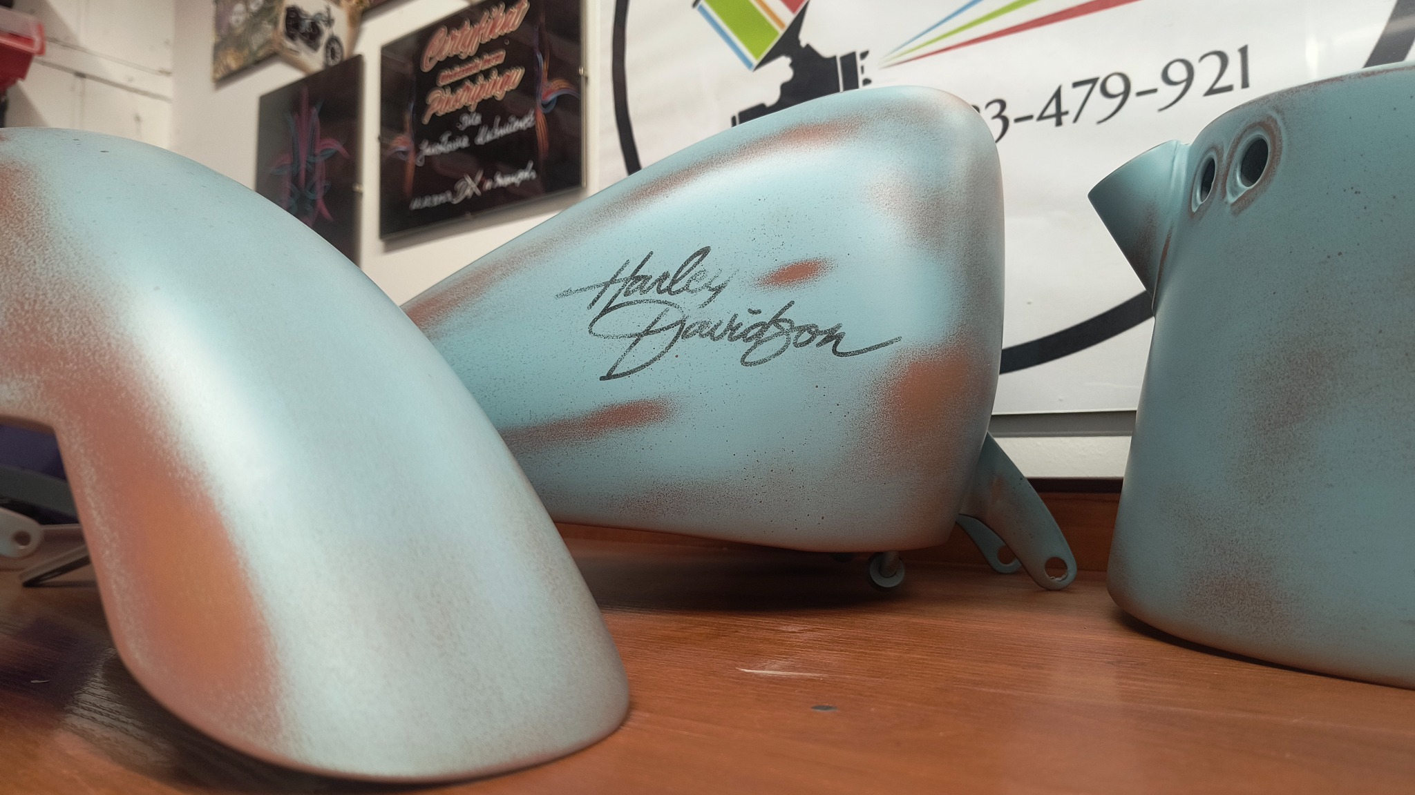 Pomalowane na niebiesko z efektem rdzy elementy motocykla Harley Davidson: błotnik, zbiornik paliwa z napisem i element ramy, leżą na drewnianym blacie.