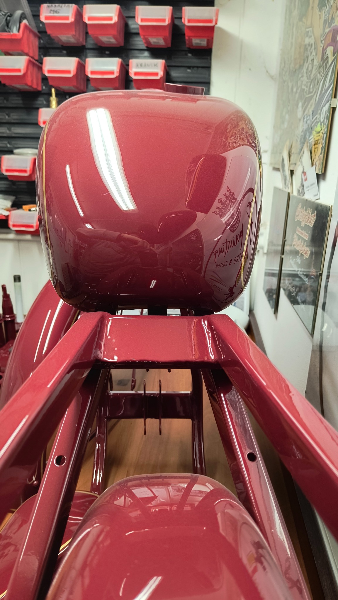 Błyszczący, bordowy bak motocyklowy z delikatnym złotym paskiem i napisem, umieszczony na metalowej konstrukcji, widoczne elementy błotnika, w tle ściana z narzędziami i grafikami.