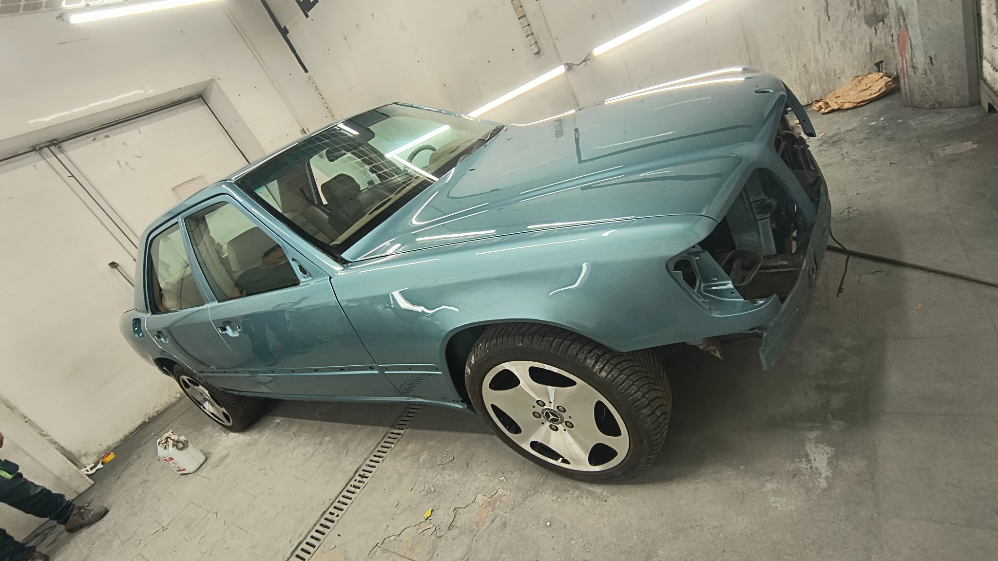 Turkusowy Mercedes W124 bez przedniego zderzaka w lakierni, po świeżym lakierowaniu. Widoczne odbicia światła na karoserii, podkreślające gładkość powłoki.