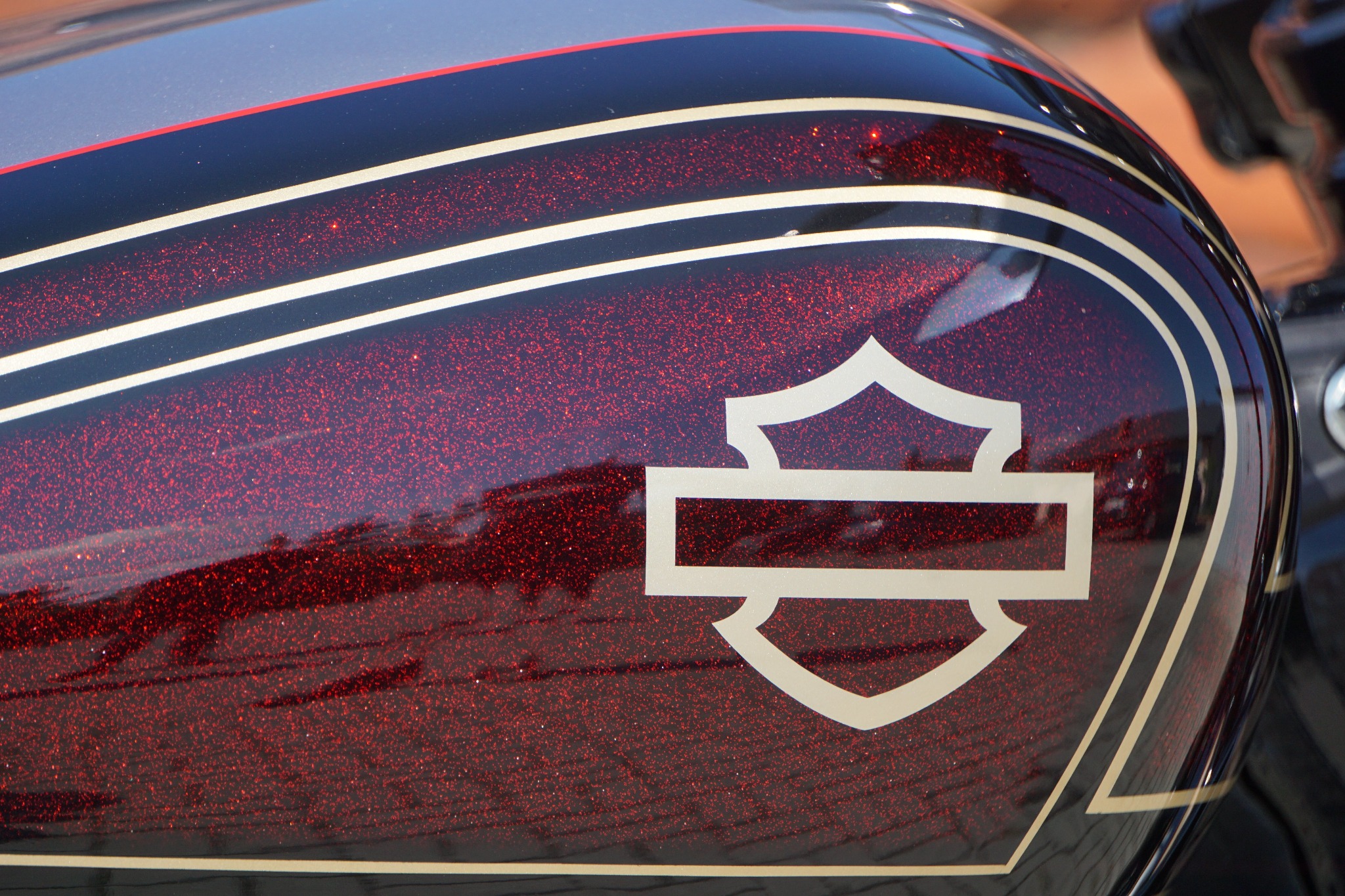 Zbliżenie na błyszczący, bordowy bak motocykla z białym logo i paskami. Widoczne refleksy światła podkreślają głębię koloru i precyzję wykonania detali.