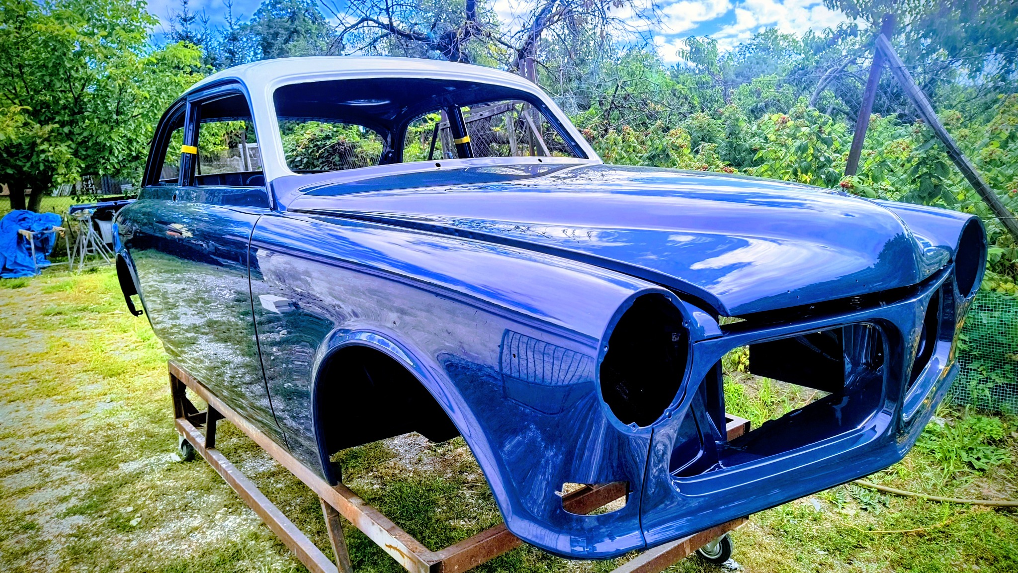 Niebieska karoseria klasycznego auta, po renowacji lakierniczej, umieszczona na metalowym stelażu na tle zieleni. Widoczne odbicia nieba i drzew w lakierze.