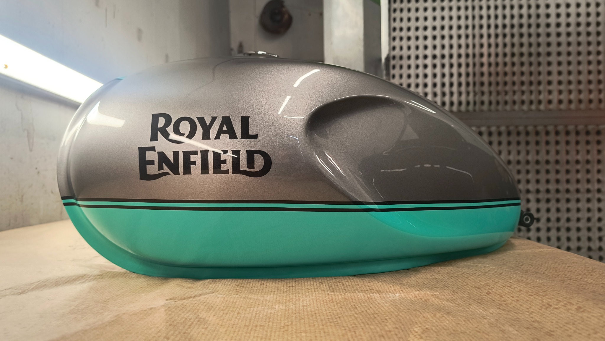 Pomalowany zbiornik paliwa motocykla Royal Enfield w kolorze szaro-turkusowym z czarnym logo, leżący na stole w warsztacie. Widoczne detale lakieru i precyzja wykonania.