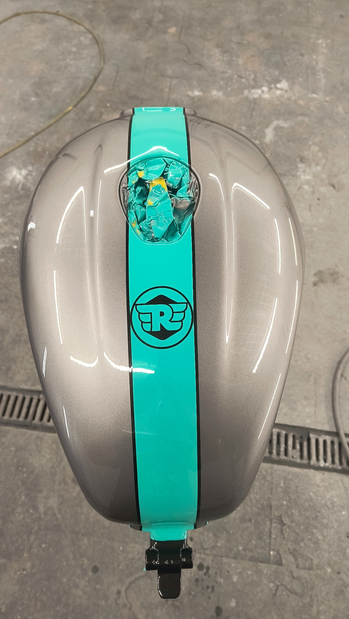 Szary bak motocyklowy z turkusowym pasem i logo, zabezpieczony folią wlewu paliwa. Połysk lakieru, widoczne odbicia światła. Detal po lakierowaniu.