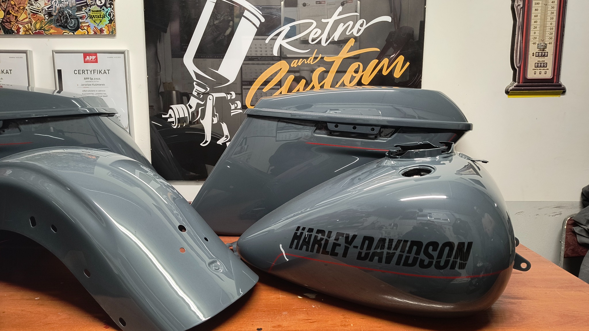 Szare, polakierowane elementy motocykla Harley-Davidson: błotnik, zbiornik paliwa i kufer, z czarnym napisem i czerwoną linią, na tle plakatu Retro and Custom.