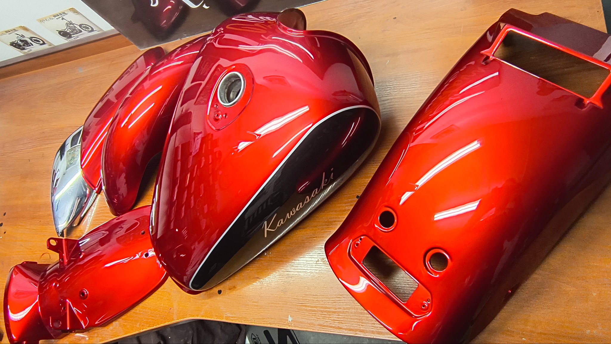 Czerwone, błyszczące elementy motocykla Kawasaki po lakierowaniu, leżące na drewnianym stole. Widoczne detale, takie jak zbiornik paliwa i błotniki.