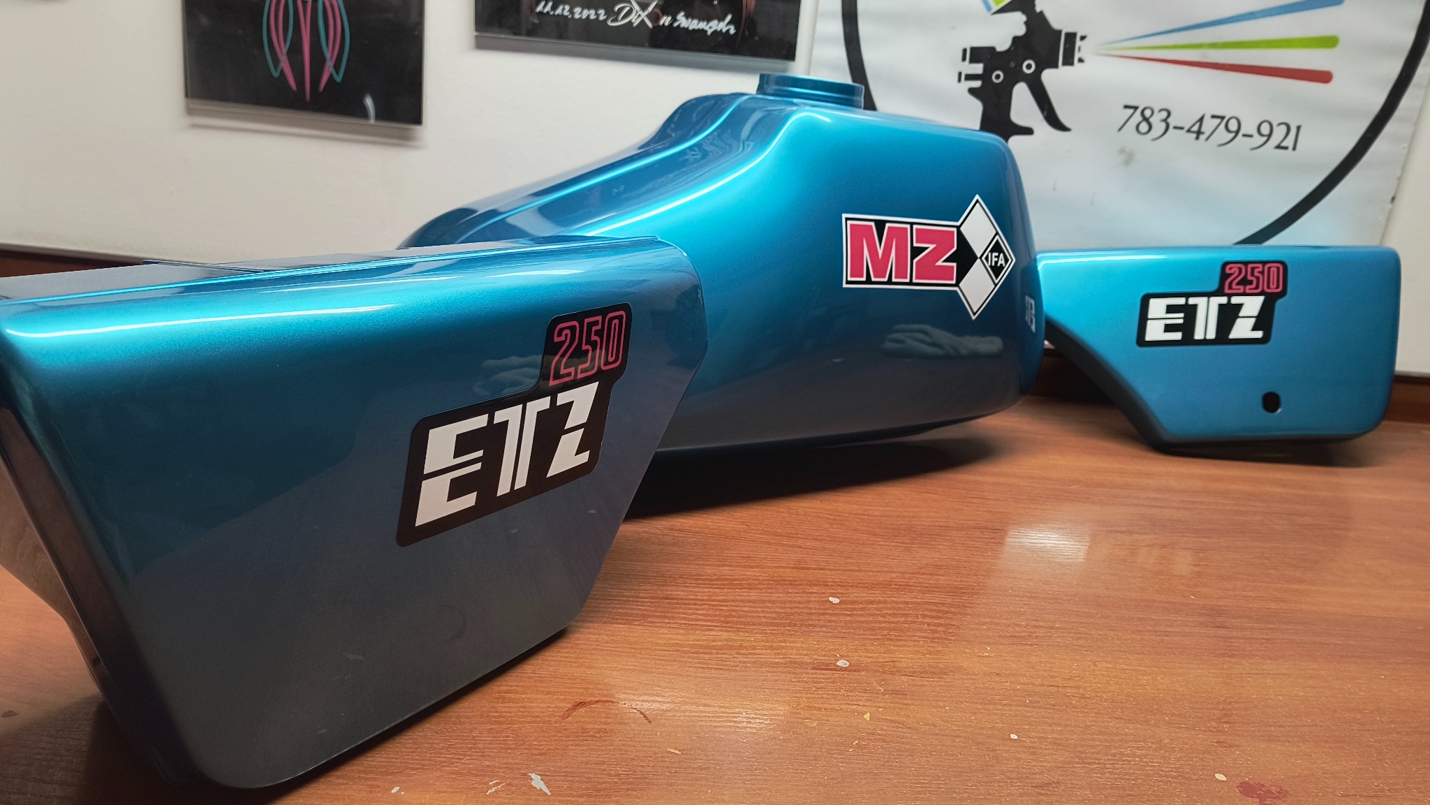 Turkusowy bak i błotniki motocykla MZ ETZ 250 po renowacji lakierniczej, z naklejkami. Błyszcząca powierzchnia, widoczne odbicia światła. Tło: ściana z grafikami.