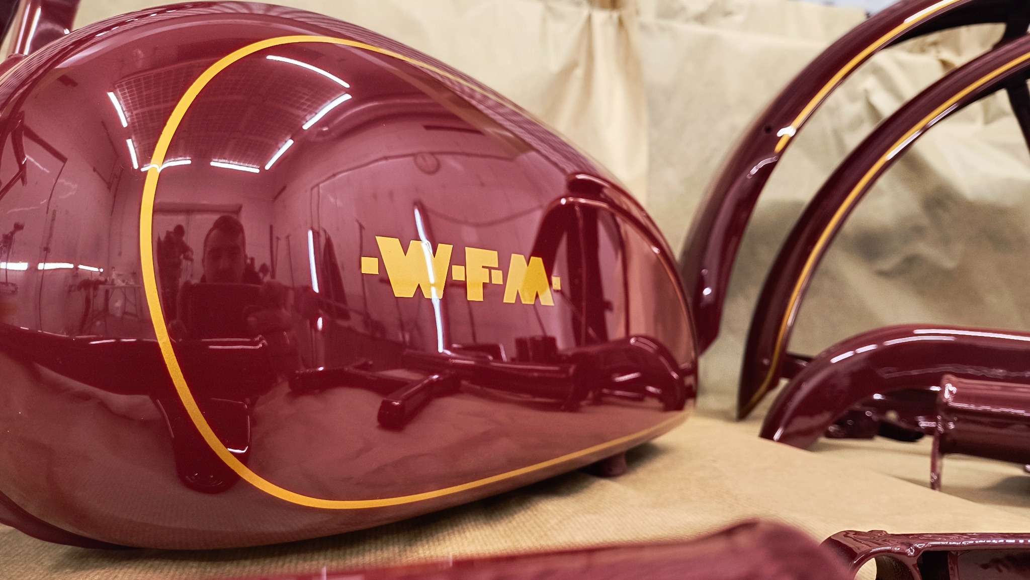 Błyszczący, bordowy bak motocyklowy WFM z żółtymi detalami i odbiciem warsztatu, obok elementy błotników po lakierowaniu. Precyzyjna renowacja.