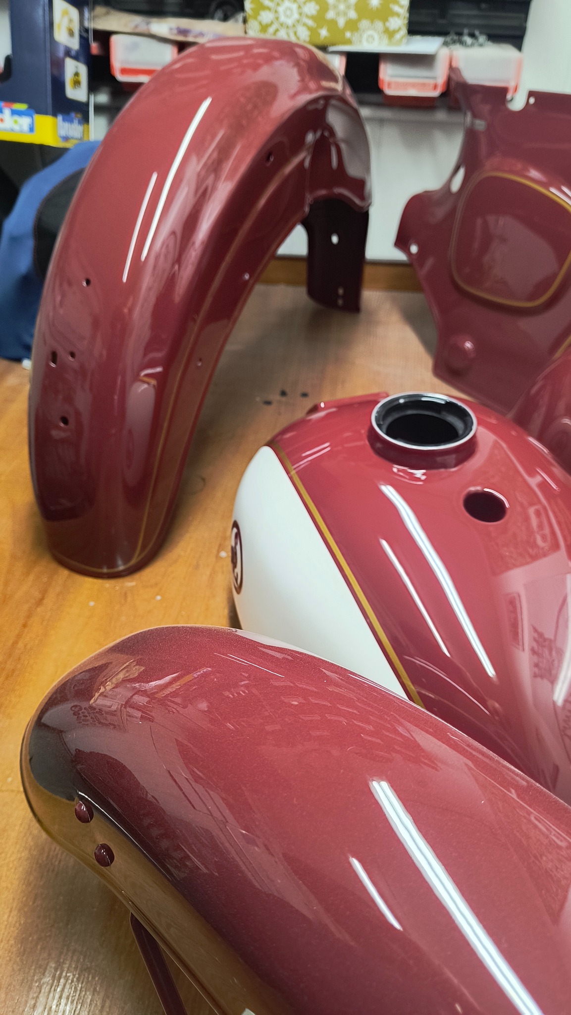 Błyszczące, bordowe elementy motocykla z białymi i złotymi akcentami po lakierowaniu, leżące na drewnianym stole. Widoczny zbiornik paliwa i błotniki.