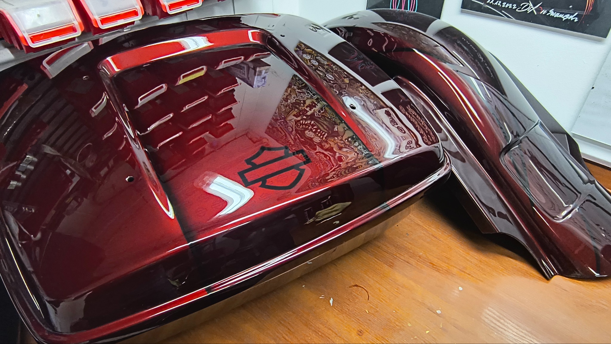 Błyszczące, bordowe elementy motocyklowe Harley-Davidson po lakierowaniu, z widocznym logo i grafiką, leżące na drewnianym blacie. Gładka powierzchnia z odbiciami.