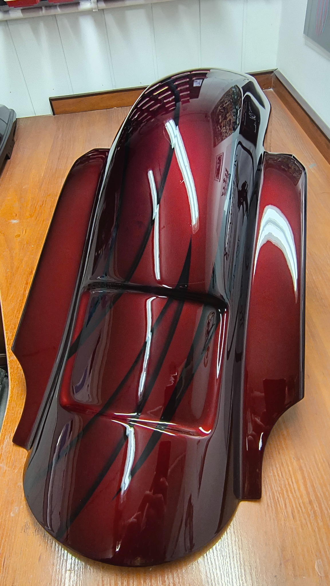 Błyszczący, bordowy błotnik motocyklowy z czarnymi, ukośnymi pasami, leżący na drewnianym stole. Widoczne odbicia światła podkreślają głębię koloru i precyzję lakierowania.