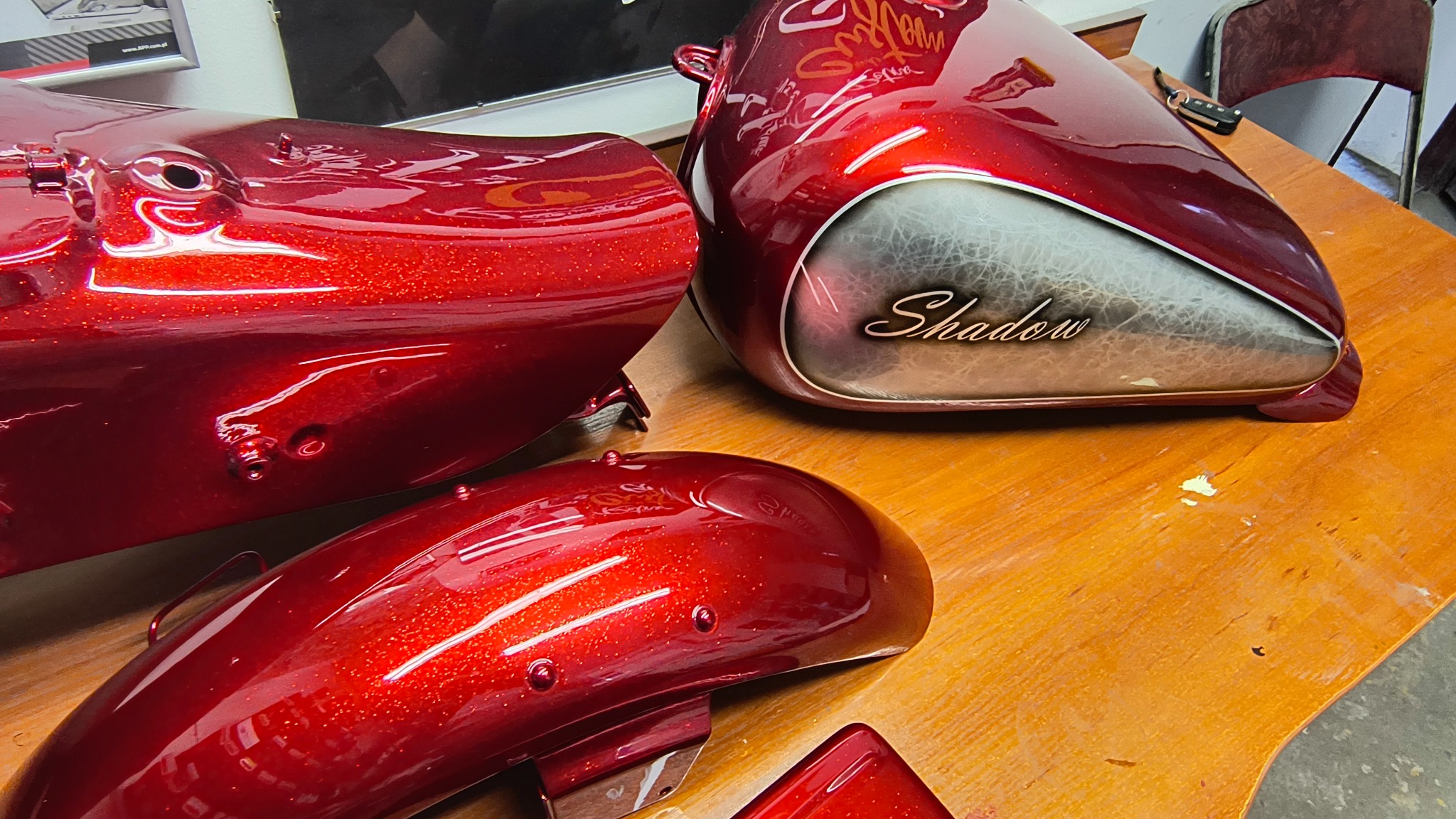 Części motocyklowe (błotnik, zbiornik) po lakierowaniu na czerwono z brokatem, zbiornik z napisem 'Shadow'. Elementy leżą na drewnianym stole.
