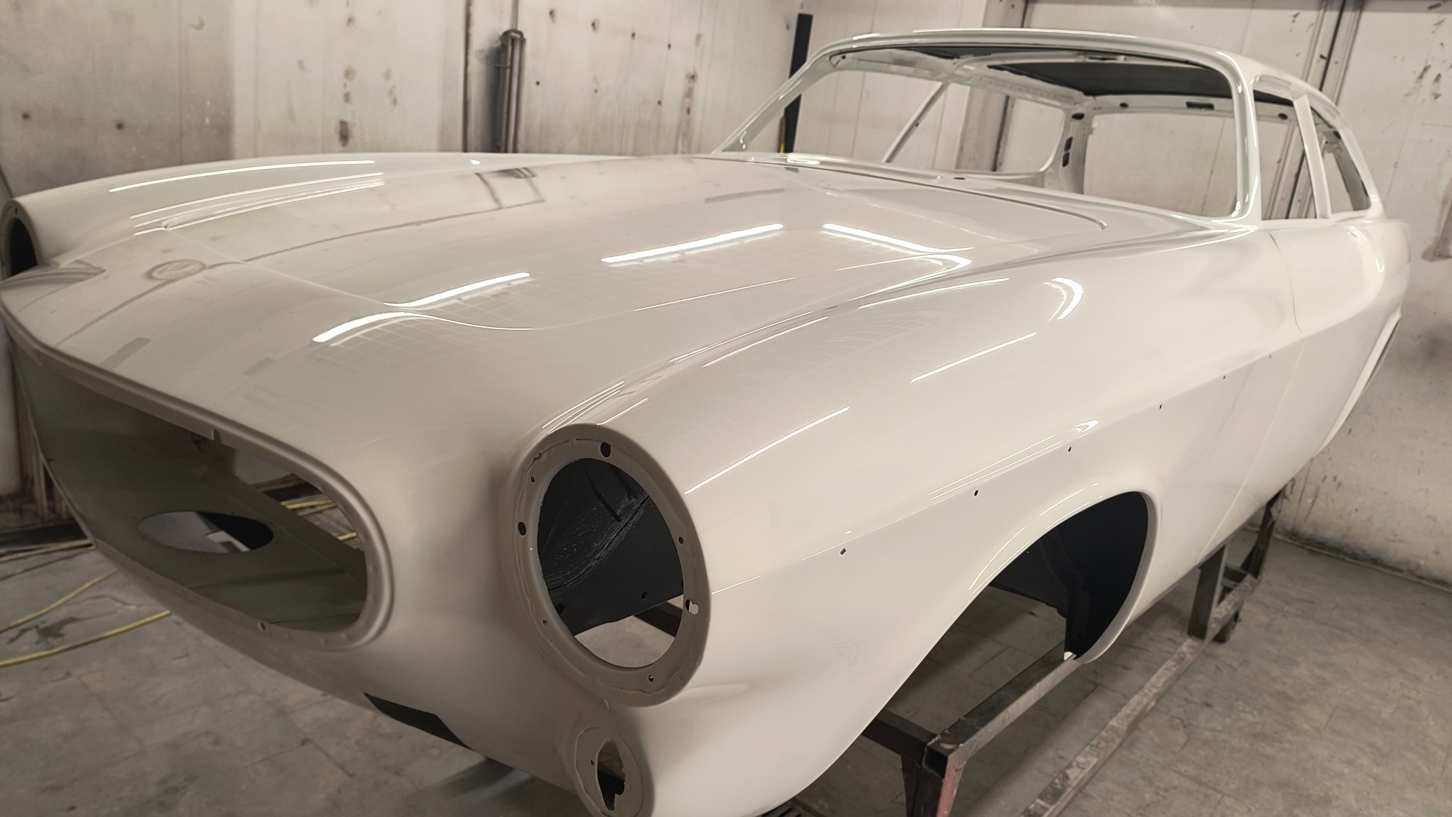 Biała karoseria klasycznego samochodu po lakierowaniu, widoczna z przodu, osadzona na metalowym stelażu w warsztacie. Gładka powierzchnia, brak elementów wyposażenia.