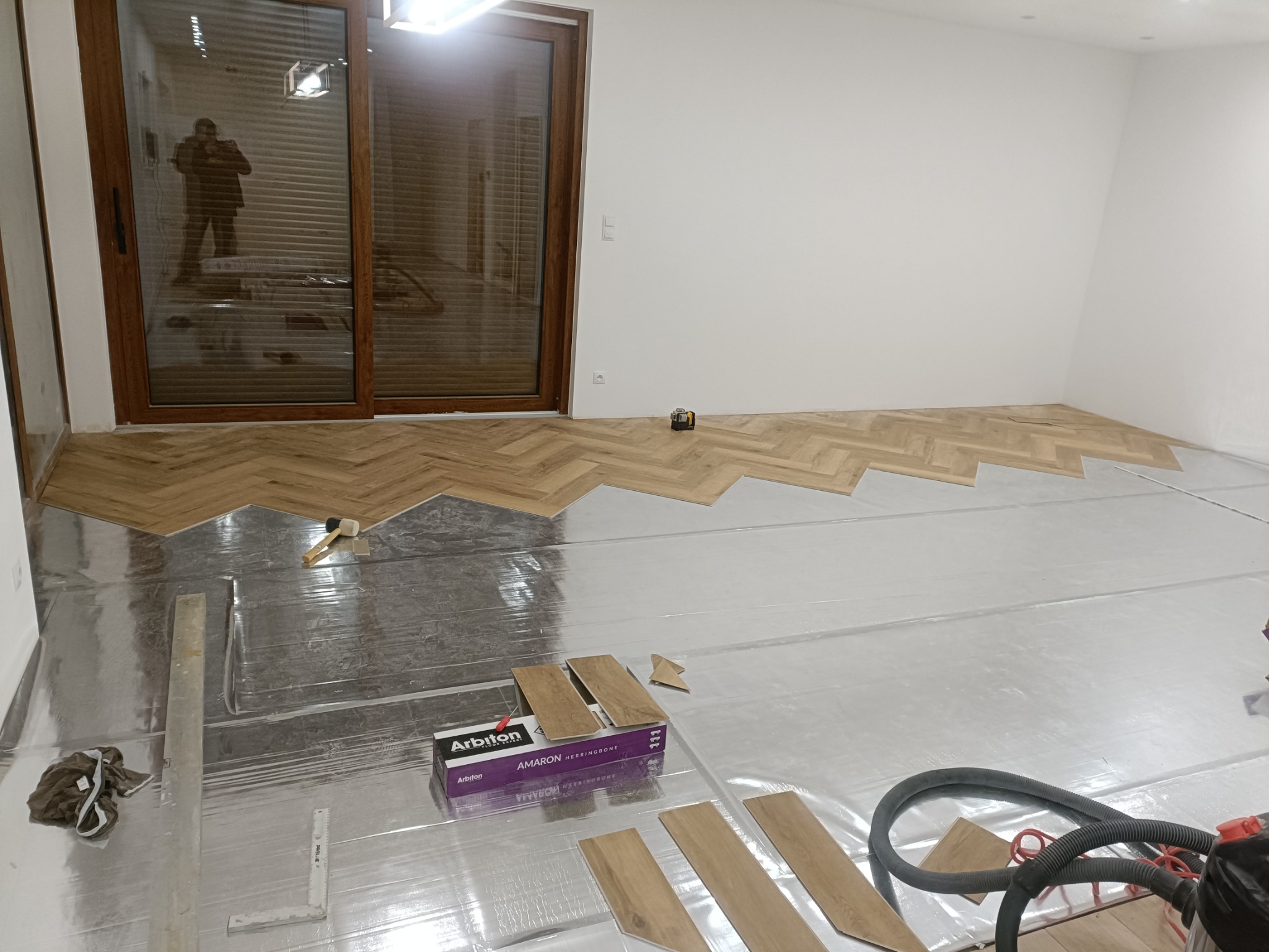 Fragment pokoju w trakcie układania podłogi w jodełkę, widoczna folia izolacyjna, częściowo ułożone panele, paczka paneli Arbiton i narzędzia pracy.