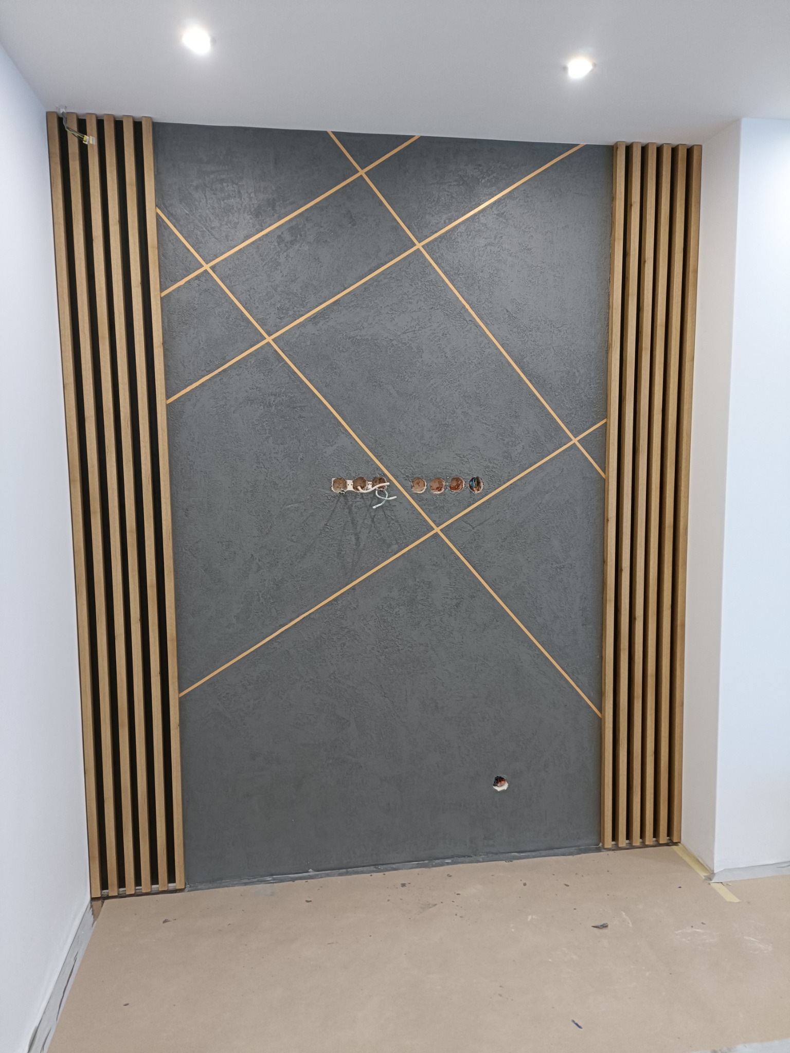 Szara ściana dekoracyjna z drewnianymi lamelami po bokach i złotymi liniami tworzącymi geometryczny wzór, widoczne puszki elektryczne bez osprzętu.