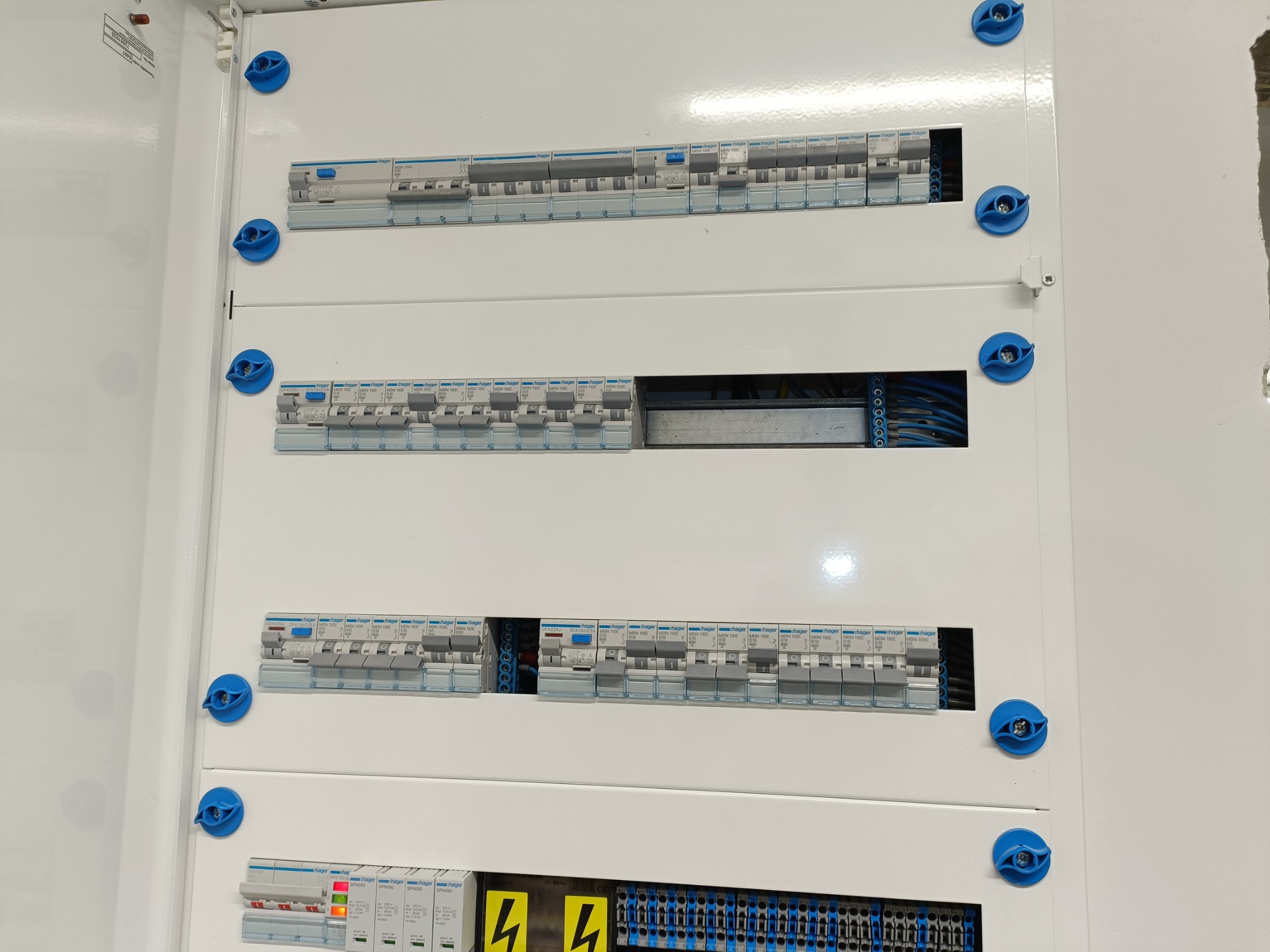 Rozdzielnica elektryczna z widocznymi bezpiecznikami i przewodami, zamknięta białymi drzwiczkami z niebieskimi pokrętłami, widok z bliska.