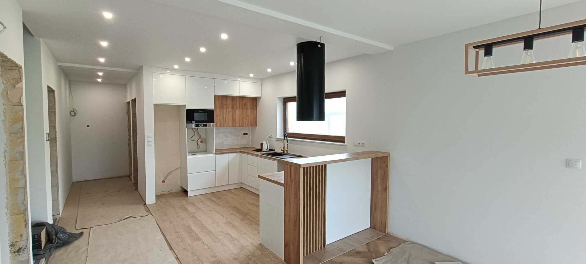 Nowoczesna kuchnia w trakcie wykończenia: białe szafki z drewnianymi akcentami, czarny okap cylindryczny, drewniany blat barowy z pionowymi listwami, podłoga w jasnym drewnie, widoczne...