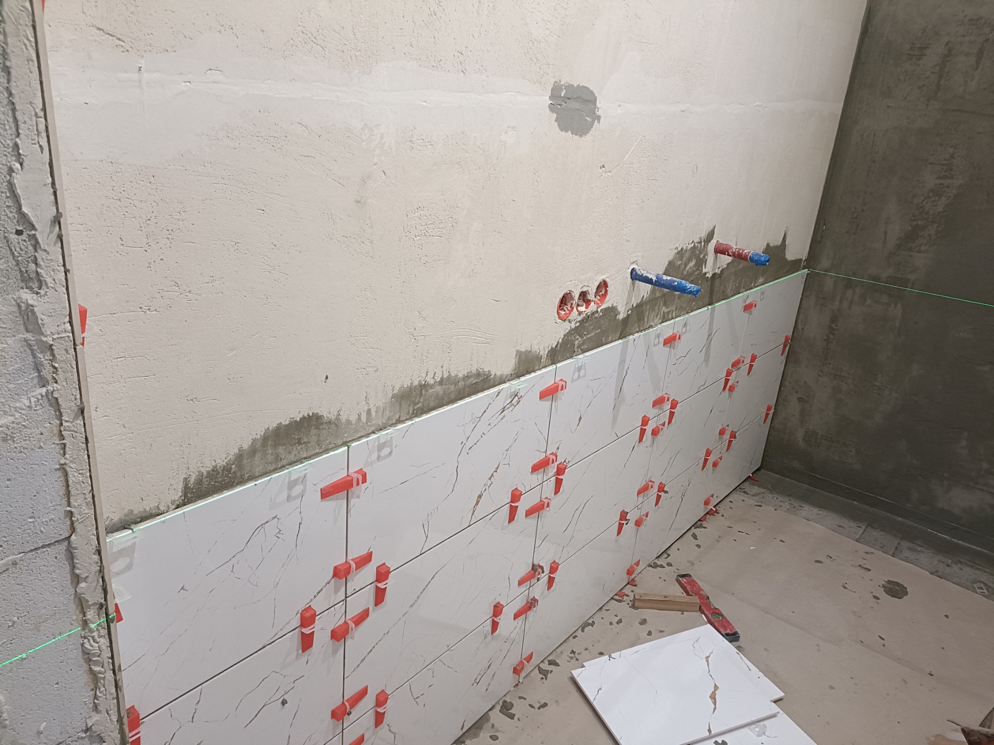 Układanie białych płytek imitujących marmur z użyciem czerwonych klipsów poziomujących; widoczne wystające rury instalacji wodnej i laserowy niwelator liniowy.