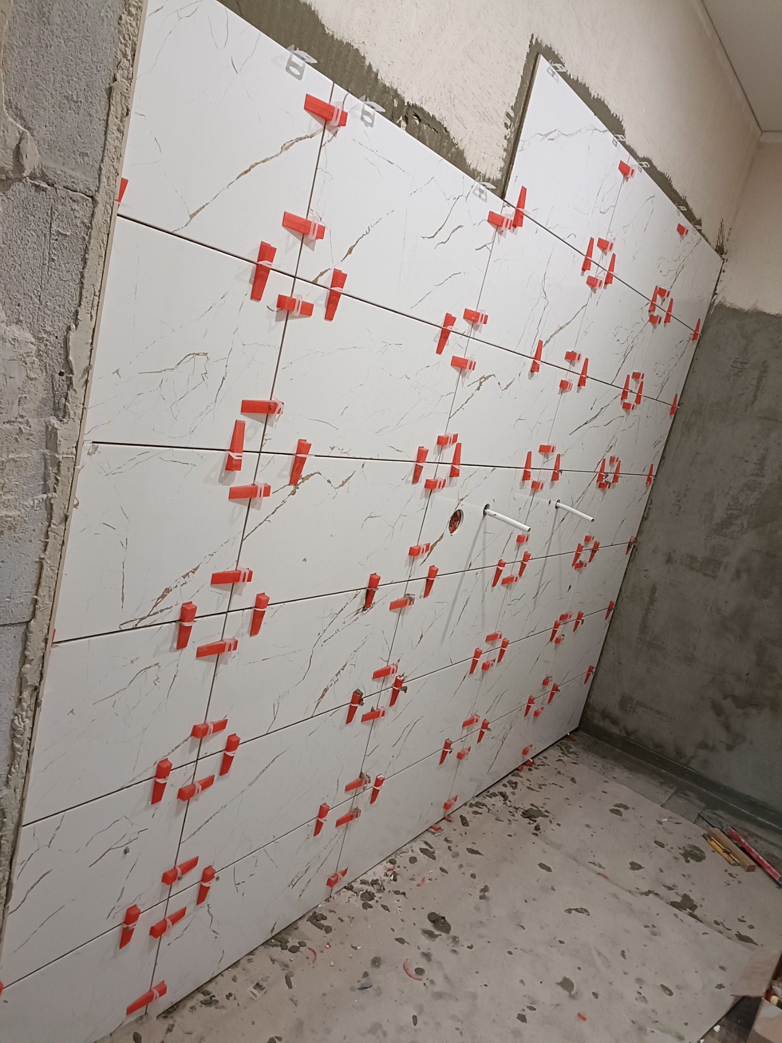 Ściana w trakcie kładzenia białych płytek imitujących marmur, z użyciem czerwonych klipsów poziomujących, widoczne fragmenty instalacji wodnej i surowy beton.