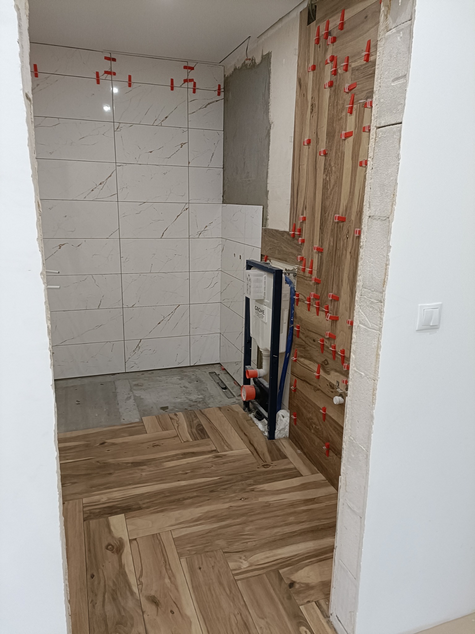 Wnętrze łazienki w trakcie remontu: białe płytki imitujące marmur na ścianie prysznica, drewnopodobne płytki układane w jodełkę na podłodze, fragment ściany z drewnopodobnych płytek z czerwonymi...