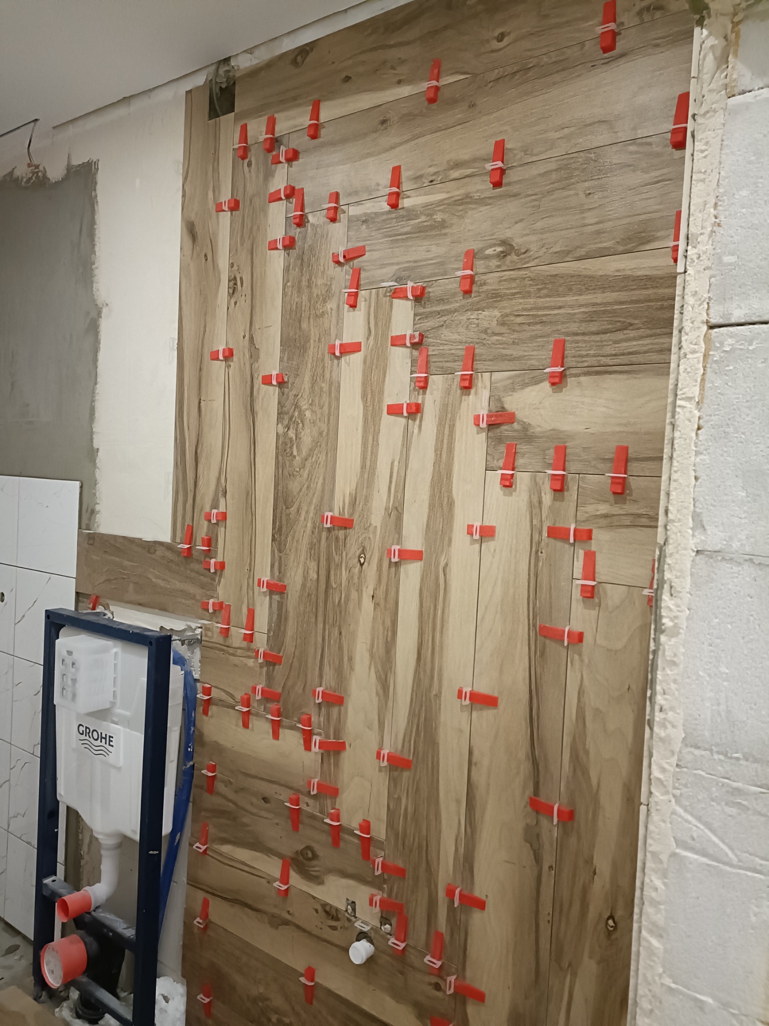 Ściana w trakcie wykańczania z drewnopodobnych płytek, z użyciem czerwonych klipsów poziomujących, widoczny stelaż podtynkowy WC marki Grohe.