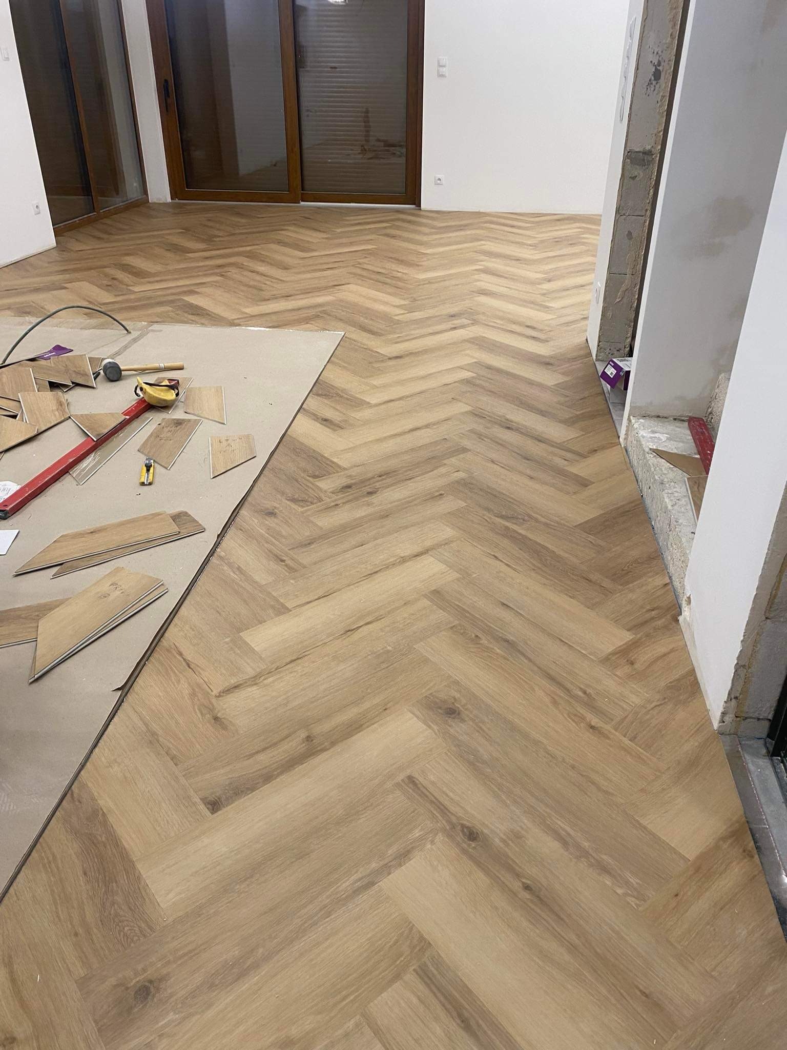 Układanie jasnych paneli podłogowych w jodełkę francuską, widoczne narzędzia i materiały na kartonie ochronnym, surowe wykończenie ścian w tle.