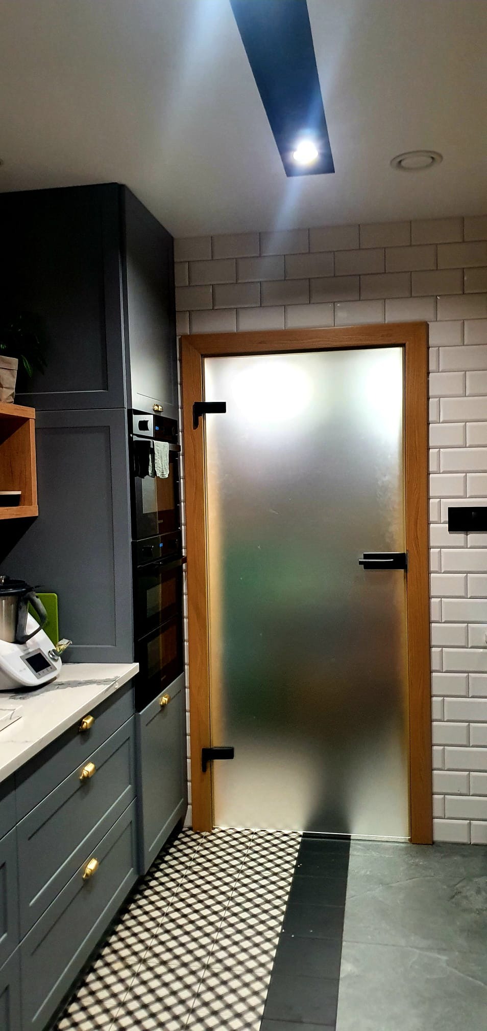 Matowe szklane drzwi z drewnianą ościeżnicą w nowoczesnej kuchni z szarymi szafkami i białą cegłą na ścianie, czarne klamki i zawiasy.