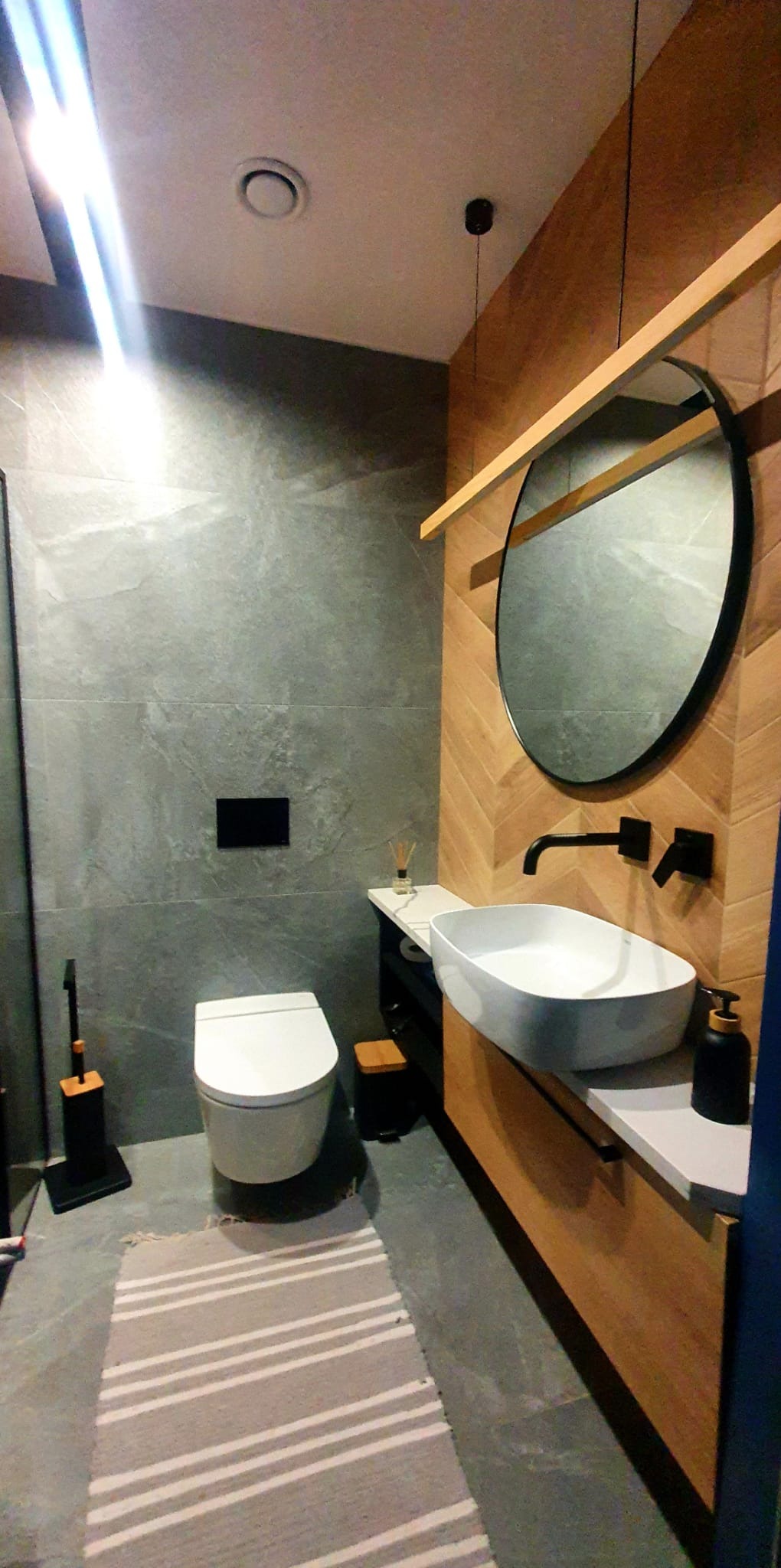 Nowoczesna łazienka z szarą płytką imitującą kamień, drewnianą ścianą w jodełkę, okrągłym lustrem, podwieszaną toaletą i umywalką nablatową.