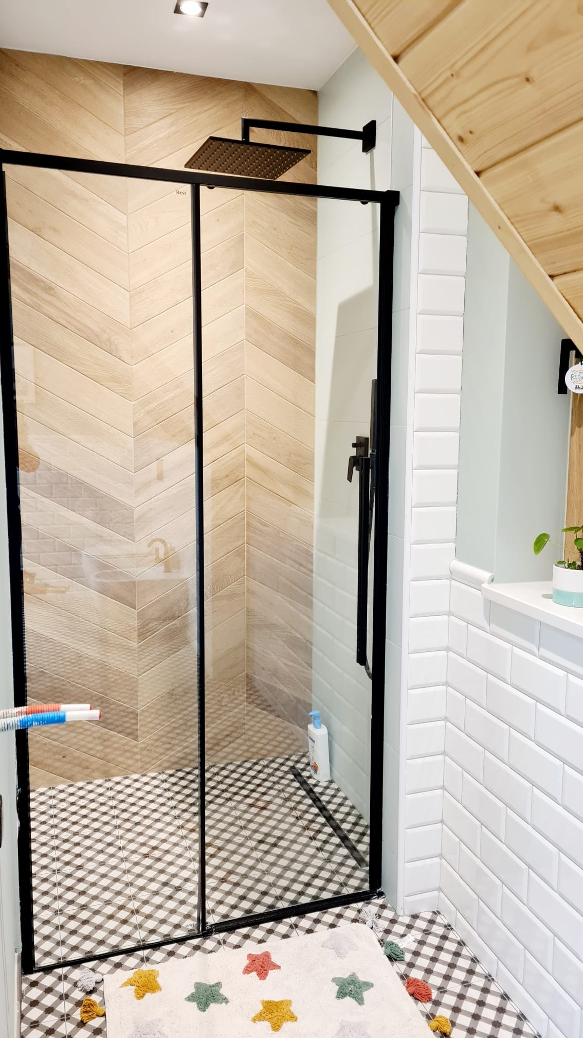 Nowoczesna kabina prysznicowa z czarnymi ramami i deszczownicą, ściany wyłożone płytkami imitującymi drewno ułożonymi w jodełkę, podłoga w czarno-białą kratę, dywanik w gwiazdki, skośny sufit...