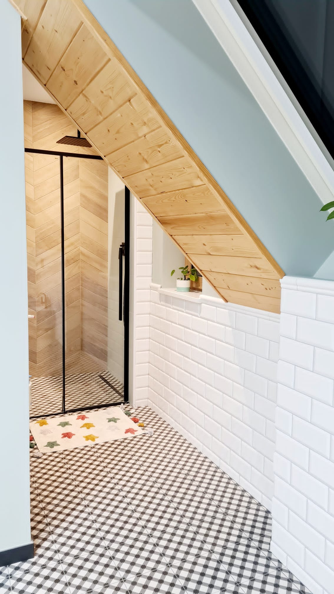 Nowoczesna łazienka na poddaszu z kabiną prysznicową w czarnej ramie, wykończona białymi płytkami typu metro i drewnianą boazerią pod skosem, z szachownicą na podłodze i małym dywanikiem przed...
