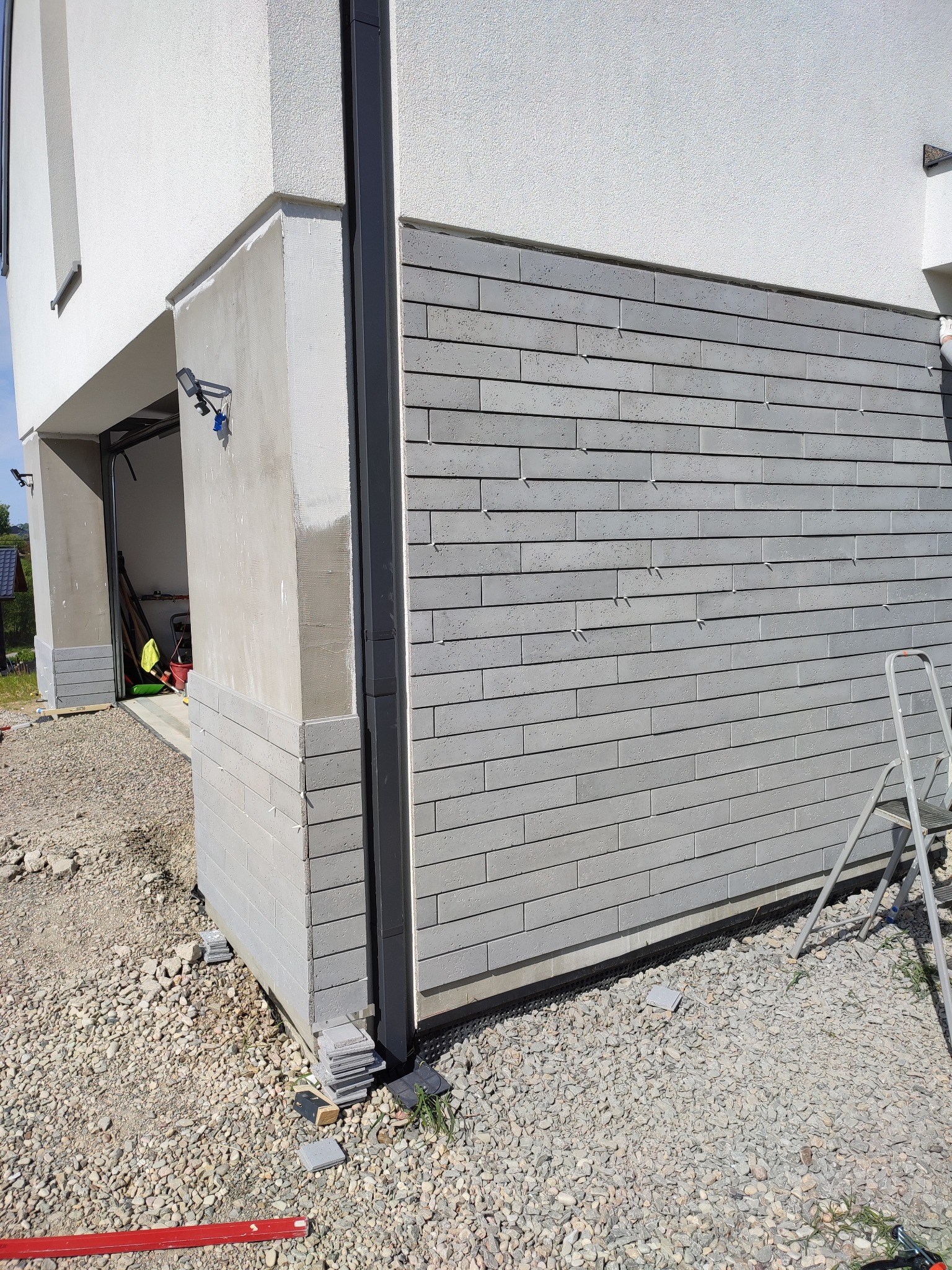 Fragment elewacji budynku z szarej cegły klinkierowej w trakcie montażu, widoczne sznurki poziomujące i drabina, teren wokół wysypany żwirem.