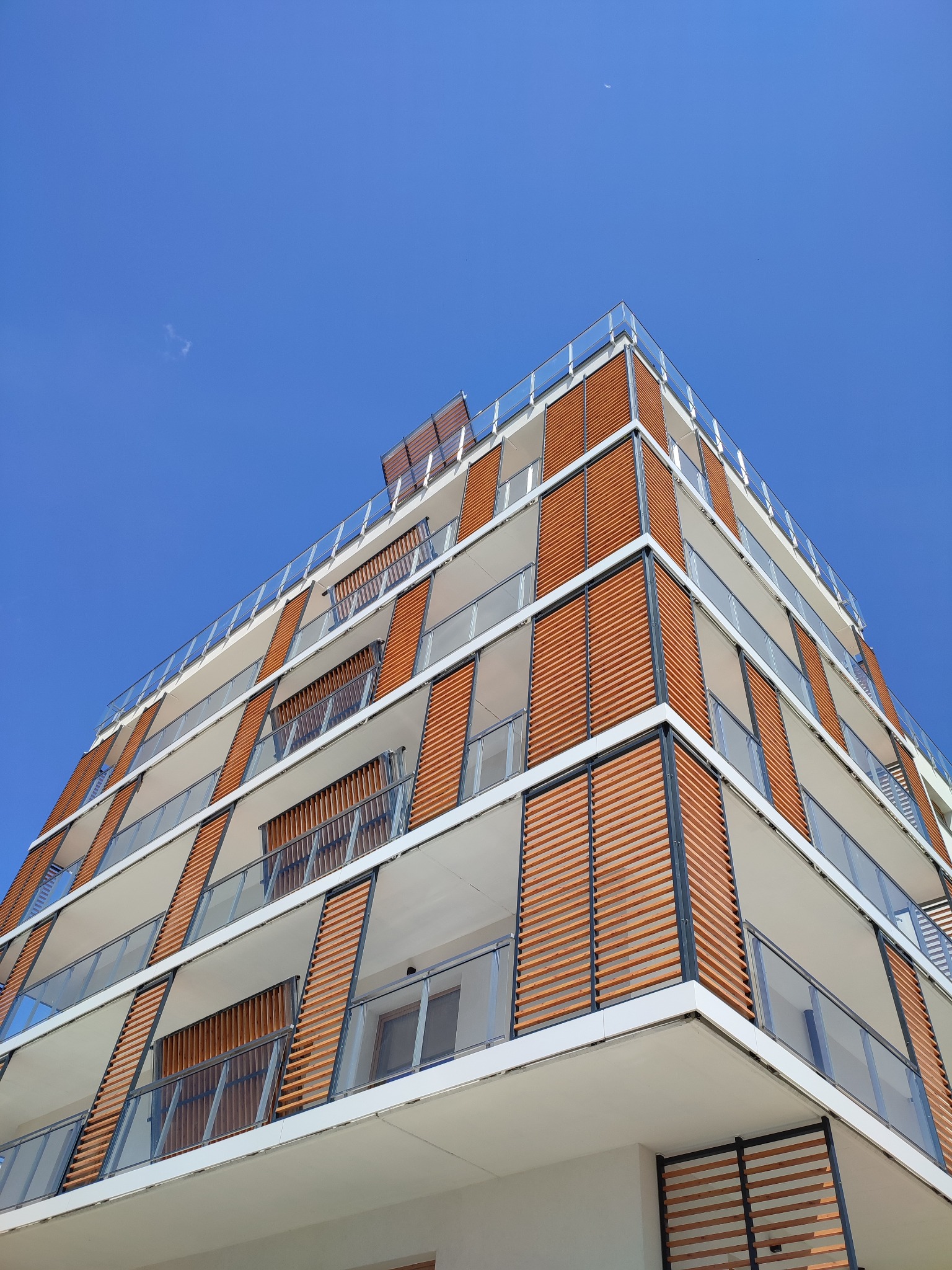 Nowoczesny budynek mieszkalny z balkonami osłoniętymi drewnianymi panelami, sfotografowany pod kątem w słoneczny dzień.