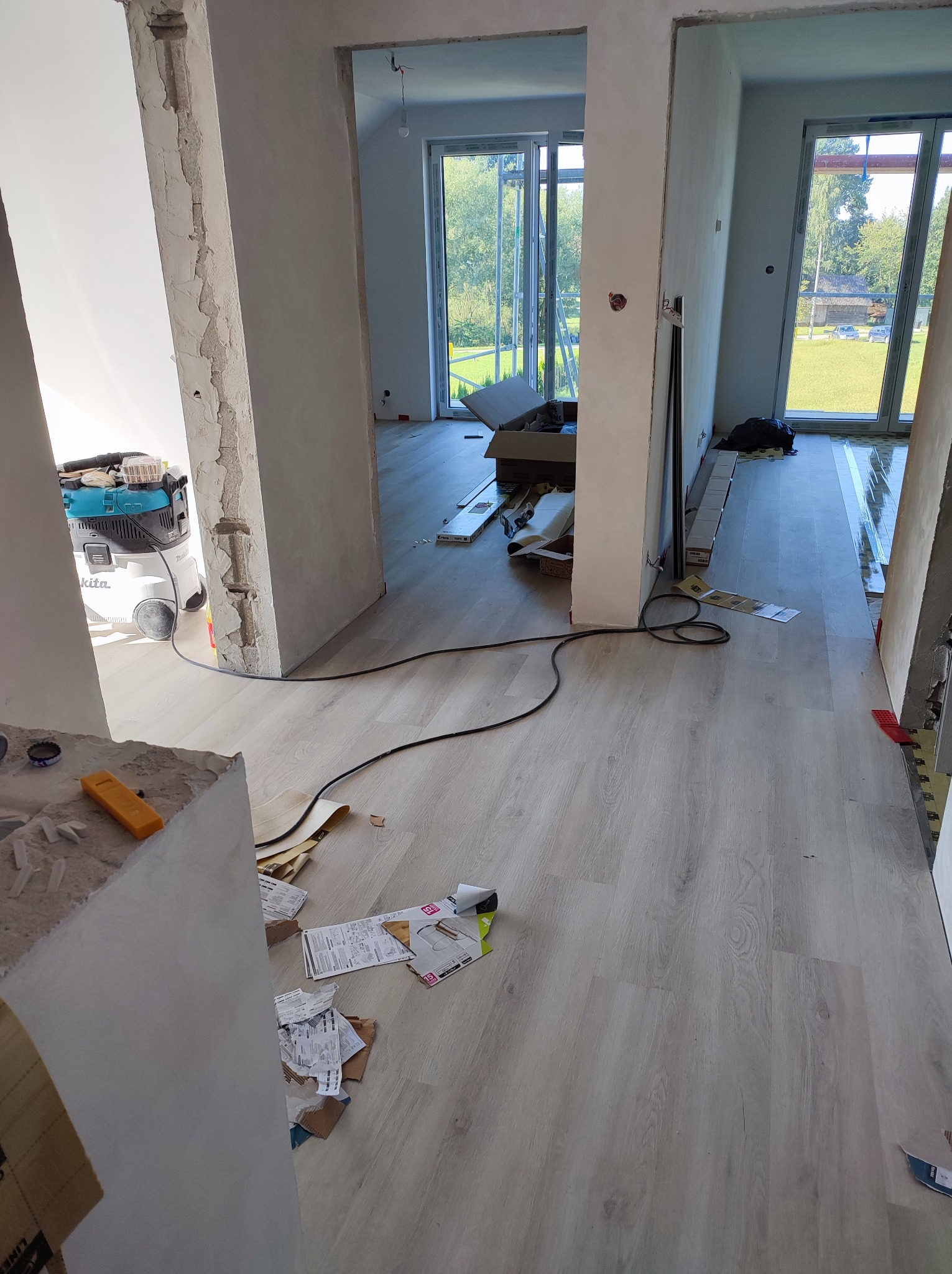 Wnętrze w trakcie remontu: ułożone panele podłogowe w odcieniach szarości, widoczne przygotowane otwory drzwiowe z obitymi framugami, resztki zaprawy, narzędzia i odkurzacz przemysłowy.