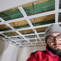 Agoos Insurance Agnieszka Przybylska - Montaż stelaża sufitu podwieszanego z widocznymi profilami, instalacją elektryczną i częściowo zamontowaną płytą gipsową, uchwycone z perspektywy wykonawcy w czerwonej polarowej kurtce i szarej...