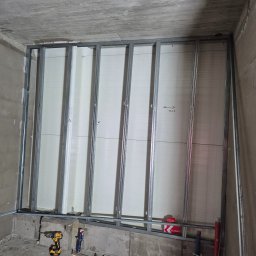 Agoos Insurance Agnieszka Przybylska - Stelaż z profili metalowych pod płytę gipsowo-kartonową przy betonowej ścianie, widoczny wkrętak akumulatorowy i opakowanie kołków montażowych.