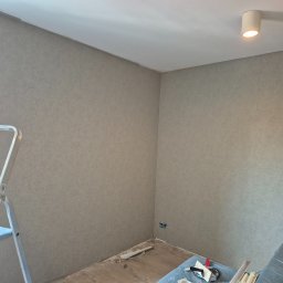 Agoos Insurance Agnieszka Przybylska - Ściany w pokoju po nałożeniu gładzi, widoczny fragment drabiny malarskiej, podłoga częściowo zabezpieczona folią, na folii narzędzia do nakładania gładzi.