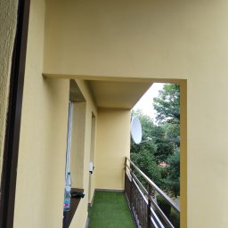 Agoos Insurance Agnieszka Przybylska - Żółty balkon z sztuczną trawą, widok na zieleń drzew i antenę satelitarną, butelka wody na parapecie.