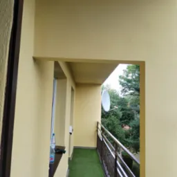 Żółty balkon z sztuczną trawą, widok na zieleń drzew i antenę satelitarną, butelka wody na parapecie.