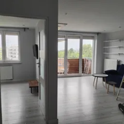 Wykończone wnętrze mieszkania w Mikołowie po remoncie, widok z perspektywy drzwi do drugiego pokoju, szare ściany, jasna podłoga, okna balkonowe, niebieska sofa i drabina malarska.