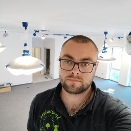 Agoos Insurance Agnieszka Przybylska - Mężczyzna w okularach, ubrany w czarną koszulkę polo z logo, robi selfie na tle pomieszczenia w trakcie prac przygotowawczych do malowania: lampy zabezpieczone folią, widoczne ślady po...