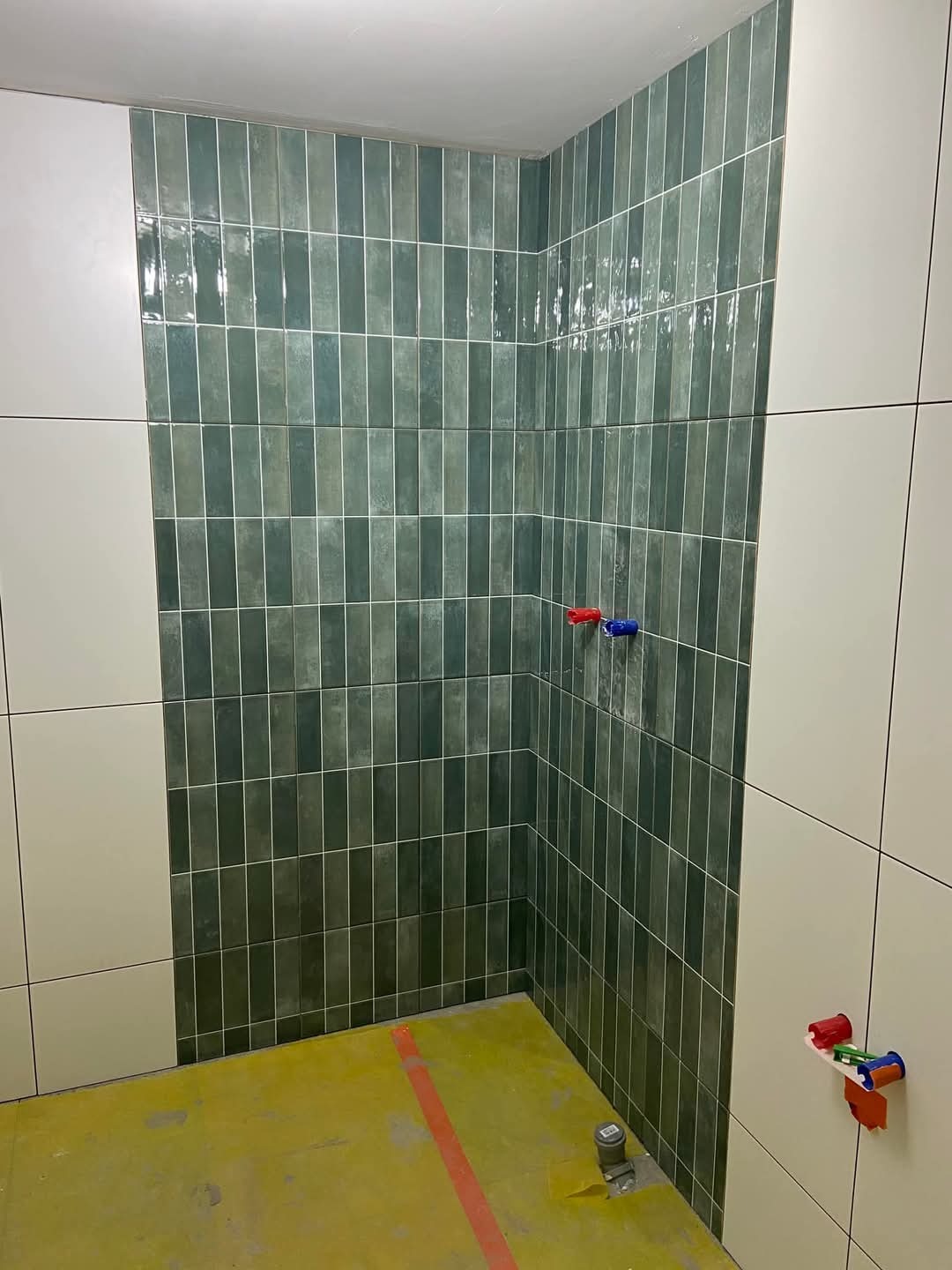 Wnętrze łazienki w trakcie prac wykończeniowych, widoczne pionowo ułożone, błyszczące, zielone płytki na ścianie, białe płytki na sąsiedniej ścianie, żółta folia ochronna na podłodze i wystające...